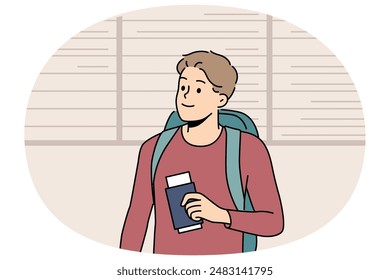 Man tourist stands at airport near board of upcoming flights and holds passport and plane ticket. Guy passenger of airport with backpack behind back is going on trip and waiting for flight