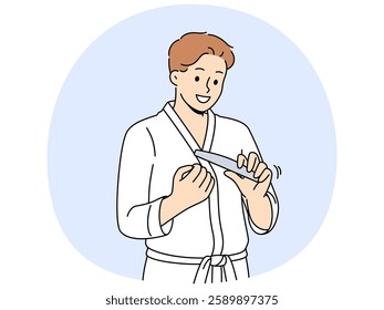 Man uses nail file after shower, performing daily hygiene and SPA procedures to take care of skin. Young cheerful guy holds nail file in hands and looks forward smiling standing in bathrobe