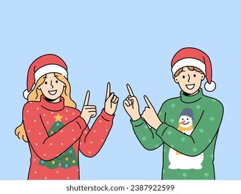 Man and woman in christmas sweaters point fingers upward to visit new year sale. Happy guy and girl in santa hats smiling looking at screen inviting you to christmas party or xmas festival