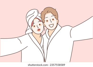 Man and woman with cosmetic masks on faces, dressed in bathrobes to perform SPA treatments. Selfie of happy couple using anti-aging clay cosmetic masks and therapeutic mud for skin tightening