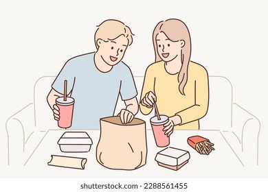 Man and woman have lunch sitting at table enjoy takeout food delivered by courier from restaurant. Guy and girl consume fast food and cold drinks, not wanting to spend time preparing dinner 