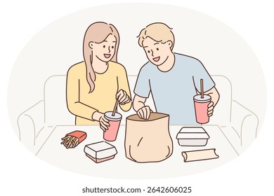 Man and woman have lunch sitting at table enjoy takeout food delivered by courier from restaurant. Guy and girl consume fast food and cold drinks, not wanting to spend time preparing dinner