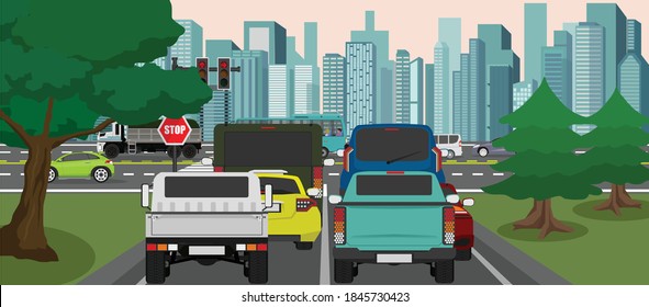 Many cars parked and queue next to a traffic signal at the intersection. Cars on the main road go back and forth. Large city scene as background. Side is full of trees and green grass.