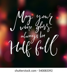 May your wine glass always be half full. Inspirational quote about wine handwritten on festive dark bokeh background with lights