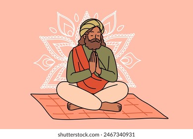 Meditating Indian man fakir prays sitting on mat with mandala sign behind back. Meditating adult guru with beard makes prayer gesture, performing esoteric and spiritualistic practices