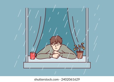 Melancholic man looks at rain from window with head on sill near coffee mug with hot drink. Melancholic guy experiences stress turning into apathy after onset of autumn due to lack of sunlight