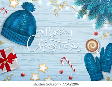 Merry Christmas Greeting Background. Winter Elements fir branches, knitted blue hat, mittens, coffee with foam, paper gift box with bow, sweets, garlands, ribbons. Hand lettering inscription. Top View