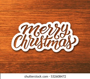 Merry Christmas text on paper label with hand lettering over wooden background. Sticker or greeting card vector design template for Xmas celebration