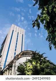 
A modern glass and concrete skyscraper towers over a weathered, curved building adorned with trees and electrical wires against a backdrop of a clear blue sky.