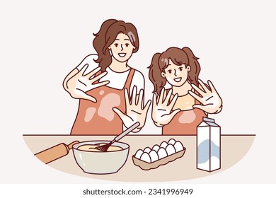 Mom and daughter are preparing breakfast or pastries and with smile show palms stained with flour. Woman brings up little girl teaching how to cook pastries from dough and enjoys raising child