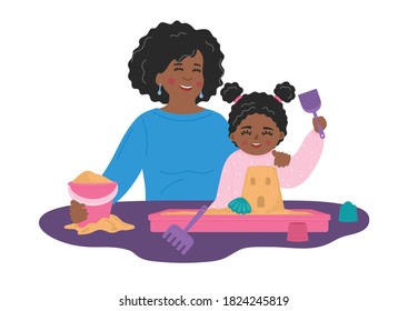 Mother with child plays with sand and toys in plastic sandbox on the table. African girl with nanny builds castle. Kids' sensory, motor skills, creativity and imagination development playing.