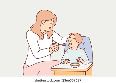 Mother feeds boy sitting in child seat with spoon, caring for baby and wanting to give son best. Woman works nanny in kindergarten and feeds baby with healthy porridge or fruit puree after midday nap