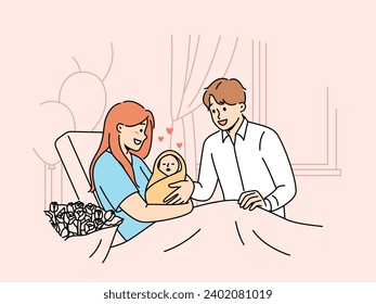 Mother with newborn baby sits on couch in hospital and shows infant to happy father. Smiling married couple have become parents for first time admire sleeping newborn wrapped in blanket