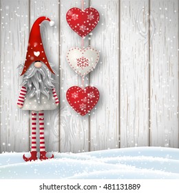 Nisser in Norway and Denmark, Tomtar in Sweden or Tonttu in Finnish, Scandinavian folklore elves, nordic christmas motif, Tomte standing in front of gray wooden wall in snow, with red decorated hearts.