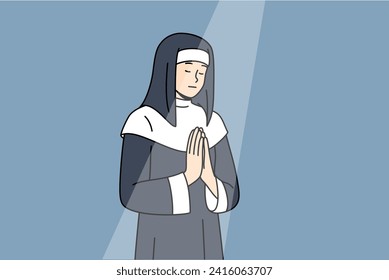 Nun prays to god with palms folded in front chest and eyes closed during ritual, standing under beam light in cathedral. Female novice catholic cathedral praying to observe christian traditions