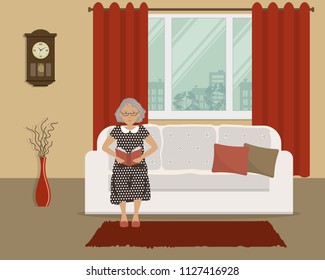 Old lady is sitting on the couch and reading a book. There is a white sofa on the window background in the living room. There is also a wall clock and a vase with decorative branches in the room