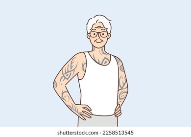 Optimistic elderly man with tattoos on arms and body stands with hands on belt proud good health in old age. Gray-haired elderly human with tattoos stuffed in youth looks at screen posing in t-shirt