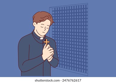 Padre prays and holds christian cross in hands, standing in confession room, asking god to atone for sins of temple visitors. Catholic priest observes ritual of confession and reads prayer