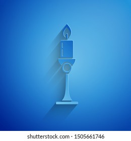 Paper cut Burning candle in candlestick icon isolated on blue background. Old fashioned lit candle. Cylindrical candle stick with burning flame. Paper art style. Vector Illustration