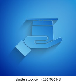 Paper cut Magician hat in hand icon isolated on blue background. Magic trick. Mystery entertainment concept. Paper art style. Vector Illustration