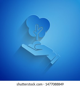 Paper cut Tree in hand of environmental protection icon isolated on blue background. Seed and seedling. Planting sapling. Ecology concept. Paper art style. Vector Illustration