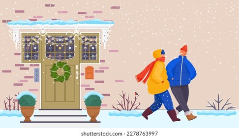 People wearing warm winter clothes walk down city street outside in cold season. Woman and man walking in snowy weather at house wall with entrance door decorated with Christmas wreath and decorations