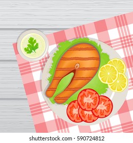 Plate. It is a fish grill on lettuce leaf. Side lined pieces of tomato and lemon. Next to the dish is the sauce on top of which are the leaves of parsley