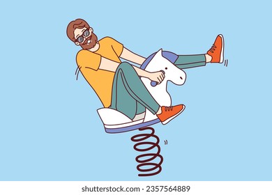 Playful man sits on wooden rocking horse and enjoys children games, behaves like child of adolescence. Bearded man in sunglasses looks at screen, is childish and does not want to grow up