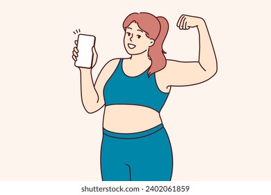 Plump woman in fitness clothes demonstrates biceps and encourages use of mobile phone with sports applications. Girl experiencing problems with excess weight does fitness to become slim and beautiful.