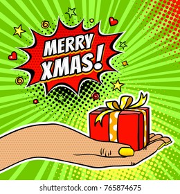 Pop art background with female hand holding a gift and Merry Christmas dynamic expression speech bubble with stars and hearts on halftone background. Vector bright illustration in retro comic style.