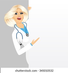 portrait of beautiful female blonde doctor peeking out of a blank presentation board and showing something on it