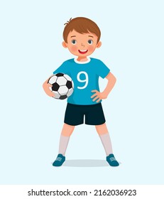 Portrait of cute little boy in sportswear holding a soccer ball posing with hand on the waist