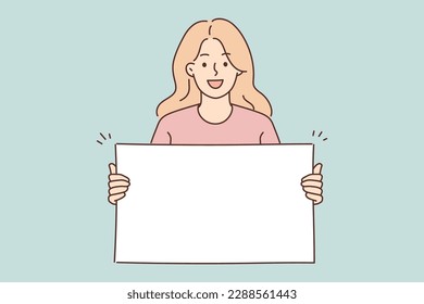 Positive woman holding blank white placard smiling and looking at camera demonstrating banner with copy space. Young girl teenager shows blank banner for presentation of promotional product 