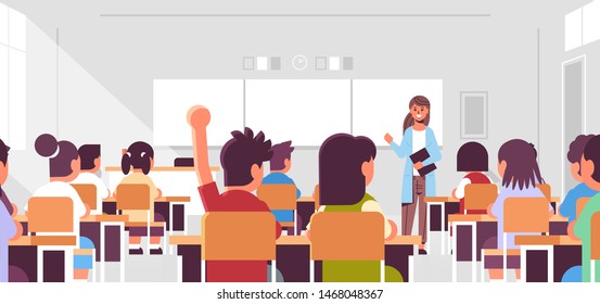 pupils group listening to female teacher schoolboy raising hand to answer in classroom during lesson teaching education concept modern class room interior flat horizontal portrait
