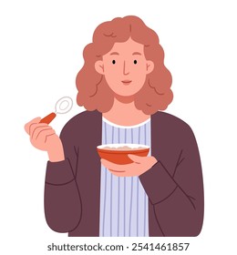 A red-haired girl in a brown cardigan eats oatmeal from a red plate and holds a spoon in her hand.The girl is eating.