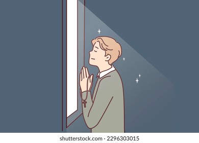 Religious man prays standing at window that emits light and holds chain with christian crucifix in hands. Believing guy prays turning to god is adherent of orthodox or catholic faith 