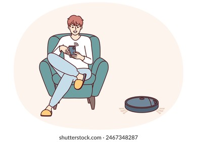 Robotic vacuum cleaner guided using mobile phone cleans floor in apartment near man sitting in chair. Robot vacuum cleaner with wi-fi chip is controlled via IOT and integrated into smart home system