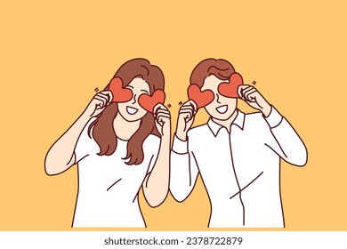 Romantic couple of man and woman holds valentine cards in form of hearts in front of eyes and looks at screen smiling. Romantic guy and girl in white clothes rejoicing in presence of soulmate