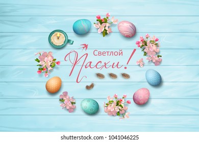 Russian inscription Happy Easter inside decorative frame. Circular border consisting of spring flowers, eggs, feathers, candle on wooden background. Top view composition