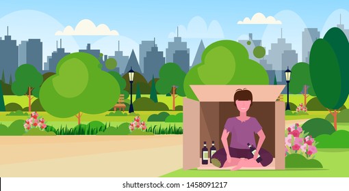 sad man drunk beggar sitting outdoor in cardboard box with bottle of alcohol homeless jobless unemployment concept city urban park cityscape background horizontal flat full length