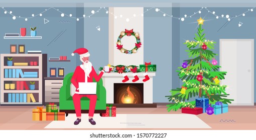 santa claus sitting in armchair using laptop and drinking tea modern living room with fireplace fir tree and gift boxes merry christmas new year holidays celebration concept horizontal full length