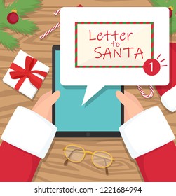 Santa claus is sitting at his workplace desk and receiving letter on his tablet - vector flat design illustration. Santa is holding his tablet on wooden desk.