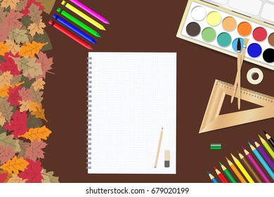 School supplies and a blank notepad ready for your text are lying on a brown wooden table. The edge forms colorful autumn leaves.