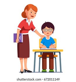 School Teacher Standing Watching, Helping, Encouraging Her Student Putting Hand On His Shoulder. Boy Kid In Glasses Sitting At The Desk And Writing In Exercise Book. Flat Vector Illustration Isolated.