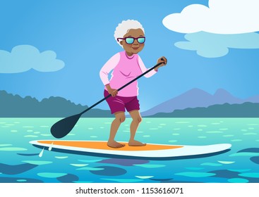 Senior African American Woman On A Stand Up Paddle Board. Cute Grandma Wearing Rash Guard And Shorts Paddling On Calm Water, Trees And Mountains In The Background. Healthy Active Lifestyle For Seniors