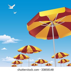 Several rows of brightly colored red and yellow beach umbrellas against brilliant blue sky with white clouds. Two seagulls are flying high above