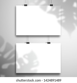The shadow of the Monstera plant falls on two white sheets of paper hanging on the clips. Mockup in horizontal orientation
