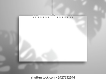 The shadow of the Monstera plant falls on a white blank spiral calendar or notebook that hangs on a thread near the wall. Realistic layout for design