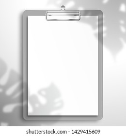 The shadow of the Monstera plant falls on gray clipboard with a blank white sheet of paper. Top view mockup template with shadow