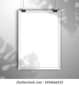 The shadow of the Monstera plant falls on blank white frame hang on clips. Mockup for design